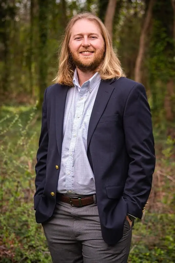 A man with long hair and beard standing in front of trees.